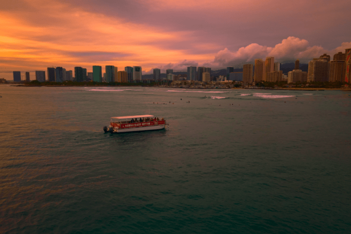Boat on ocean at sunset near city skyline with cloudy sky.