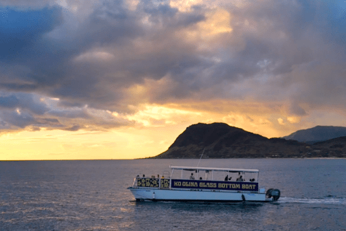 Boat on ocean at sunset with cloudy sky and distant hills.